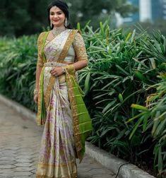 a woman in a white and green sari standing next to some plants with her hands on her hips