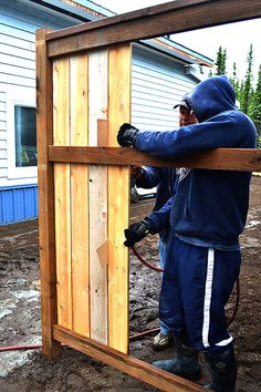 two men working on a wooden structure with hoses attached to the side and one man in blue hoodie standing next to it