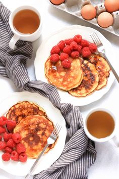 pancakes with raspberries and syrup on white plates next to cups of tea, eggs and utensils