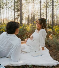 a pregnant woman sitting on a blanket in the woods talking to her husband, who is wearing a white shirt and jeans