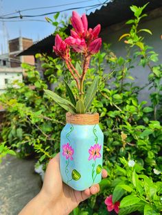 a hand holding up a painted mason jar with flowers in it and greenery behind