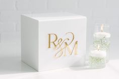 a white box with gold foiled initials and two votive candles in it next to a glass vase filled with greenery