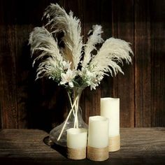 three white candles are sitting on a table with some flowers in the vase and feathers