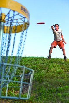 a man is throwing a frisbee in the air on a grassy hill near a disc golf basket