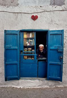 a man sitting in the doorway of a building with blue shutters and a heart shaped window