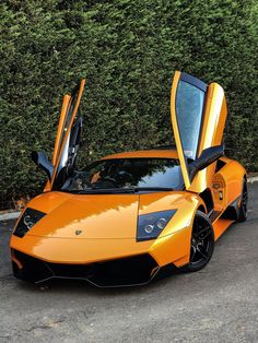 an orange sports car with its doors open in front of some bushes and shrubbery