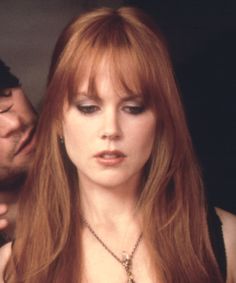 a woman with long red hair standing next to a man wearing a black shirt and necklace