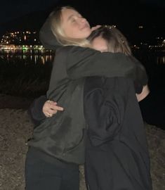 two women hugging each other while standing on the beach at night with city lights in the background