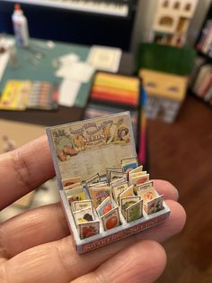 a person holding up a miniature box with stamps on it's sides in front of bookshelves