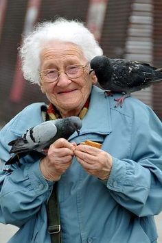 an elderly woman holding two birds on her shoulder and eating donuts in front of her