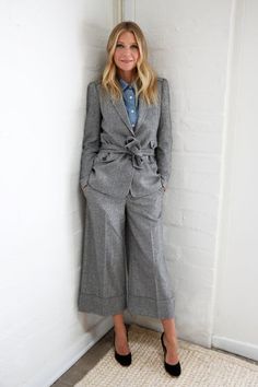 a woman standing in front of a white wall wearing a gray suit and blue shirt