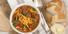 a bowl of soup with meat, beans and cheese on a cutting board next to bread