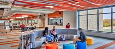 people are sitting on benches in an open area with large windows and colorful stools