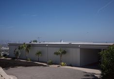 an industrial building with palm trees in the foreground and a cityscape in the background