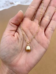 a hand holding a gold necklace with a pearl on it's chain and the ocean in the background