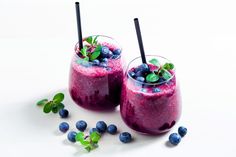 two glasses filled with blueberry smoothie on top of a white table next to fresh blueberries