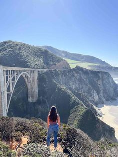 a woman standing on the edge of a cliff looking out at an ocean and bridge