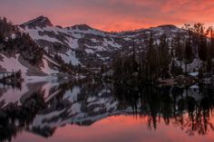 the sun is setting over a mountain range with trees in front of it and a body of water surrounded by snow covered mountains