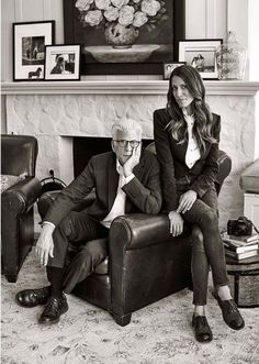 a man and woman sitting on a chair in front of a fire place with pictures above them