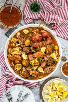 an overhead view of a bowl of stew with corn on the cob, lemon wedges and sauce