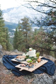 a table topped with a cake on top of a wooden cutting board next to trees