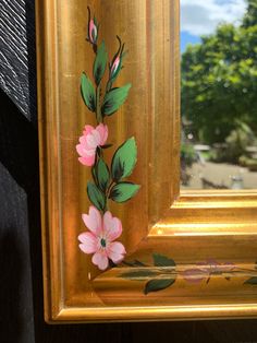 a gold framed painting with pink flowers painted on it's side and green leaves