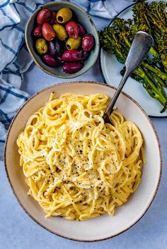 a bowl of pasta with olives and asparagus on the side next to a plate of broccoli