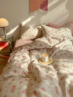 an unmade bed with pink and white flowers on it, next to a lamp