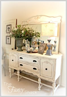 a white dresser sitting next to a mirror and vase with flowers on top of it