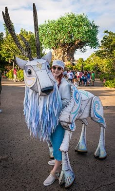 a woman in white and blue outfit standing next to a statue of a horse with feathers on it's head