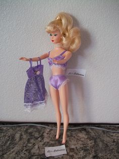 a barbie doll holding a purple bra and panties in front of a white wall with a name tag on it