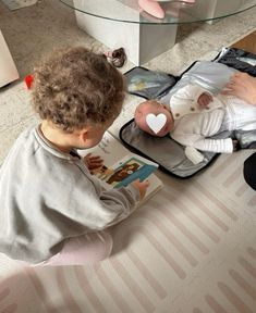 a little boy sitting on the floor reading a book to a baby doll in a suitcase