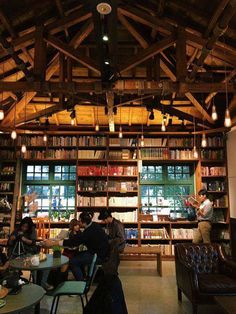 people sitting at tables in a library with lots of bookshelves and lights hanging from the ceiling