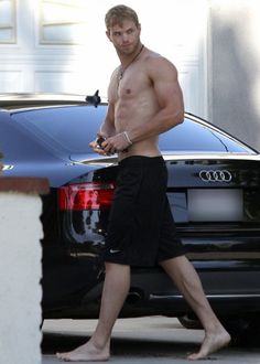 a shirtless man walking past a black car