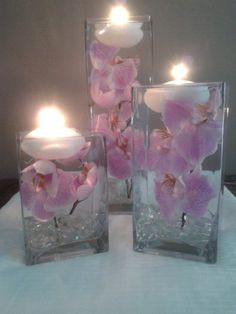 three clear vases with pink flowers in them on a white table cloth and lit candles