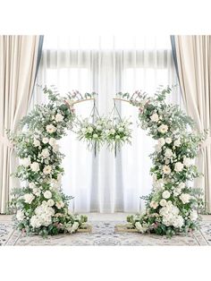 two white flowers and greenery are arranged in front of a window with drapes