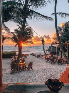 the sun is setting on the beach with chairs and palm trees in front of it