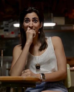 a woman sitting at a table with her hand on her mouth and looking up to the camera