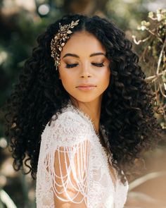 a woman with curly hair wearing a white dress and gold headpiece, standing in front of trees