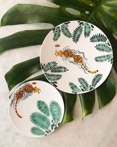 two plates with tiger designs on them sitting next to green leafy plants and leaves