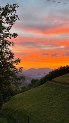 an orange and pink sunset over the mountains with trees in the foreground on a grassy hill