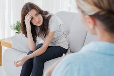 a woman sitting on a white couch talking to another woman who is holding her hand near her face