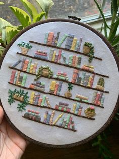 someone is holding up a cross - stitched book shelf with books on it and plants in the background