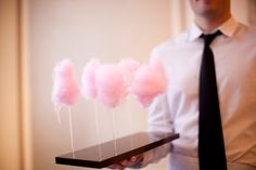 a man holding a tray with pink cotton candy on sticks in his left hand while wearing a white shirt and black tie