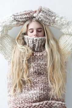 a woman with long blonde hair covering her face from the wind wearing a knitted sweater