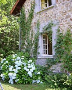 an old stone house with white windows and flowers in the foreground, surrounded by greenery