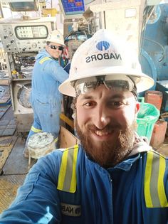 a man with a beard wearing a hard hat and safety vest in front of machinery