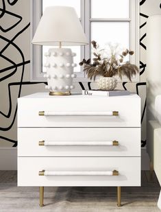 a white lamp sitting on top of a gray dresser next to a window with black and white wallpaper