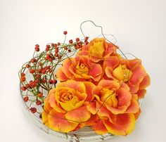 an arrangement of orange and yellow flowers in a glass bowl on a white surface with red berries