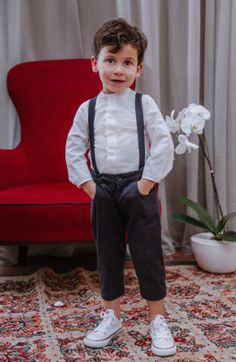 a little boy standing in front of a red chair wearing suspenders and a white shirt